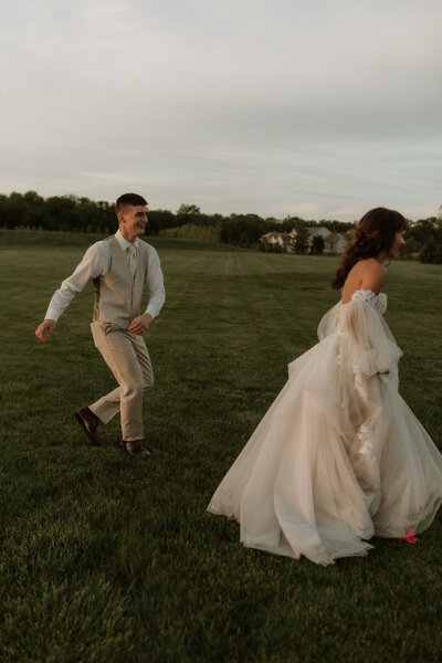 Bride and groom running together across a sunlit field at The Paddock in Fort Wayne, Indiana, from Ben and Madison’s wedding, captured by Rainstorm Photo & Video with warm, candid emotion.