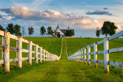 A picturesque scene of a country road flanked by white fences leading to a house on a hill. The sun casts a warm glow on the lush green grass and scattered trees, while fluffy clouds dot the blue sky in the background.