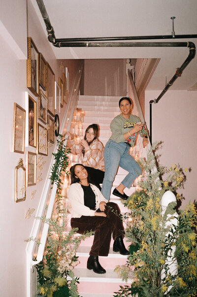 A film photograph capturing a vibrant moment during a branding session for a wedding planner. The image features three women posing playfully on a beautifully decorated staircase adorned with lush greenery, fairy lights, and framed art. Their smiles and relaxed poses reflect a joyful and approachable energy, perfectly aligning with the elegance and creativity of the wedding planner’s brand.