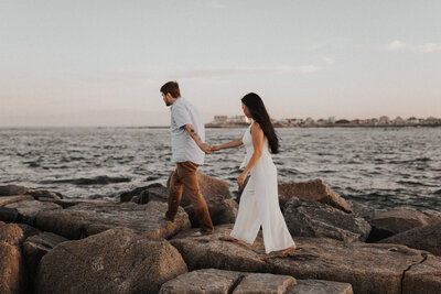 Engaged couple walks along breaker rocks next to the ocean during summer sunset
