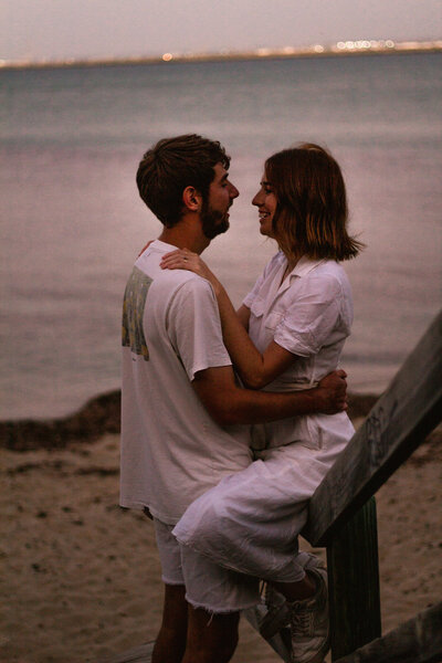 couple close together, woman sitting on wooden stair rail and man standing between her legs holding onto each other with beach in the background