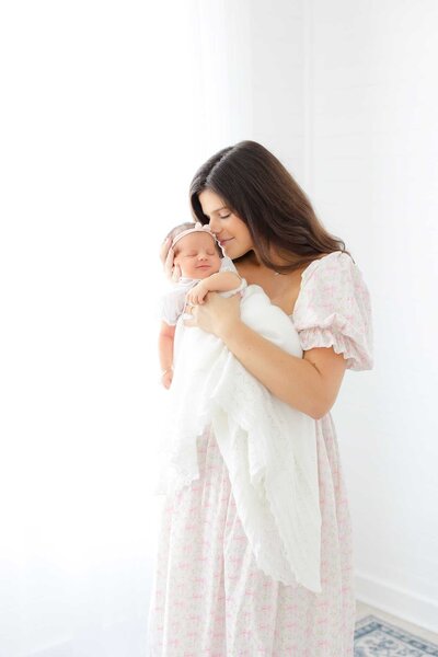 Young mom wearing a pink dress from CCP's client closet holding her newborn baby boy wrapped in a white and blue bow swaddle. She is looking at him with eyes full of love