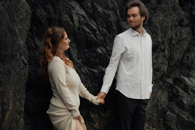 Proposal session at incinerator rock in Tofino. Couple looking at each other longingly in front of the west coasts black rock