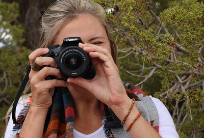 Blonde haired girl outside holding camera to eye, taking photo straight on. She's wearing a multi-color tribal scarf and a white shirt.