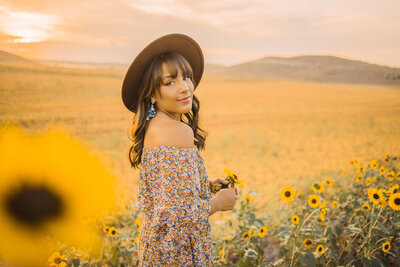 senior girl in a wide brimmed brown hat standing in a field of sunflowers