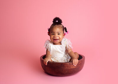Baby girl, sitting in brown bowl wearing white dress during cake smash photoshoot in Franklin Tennessee photography studio