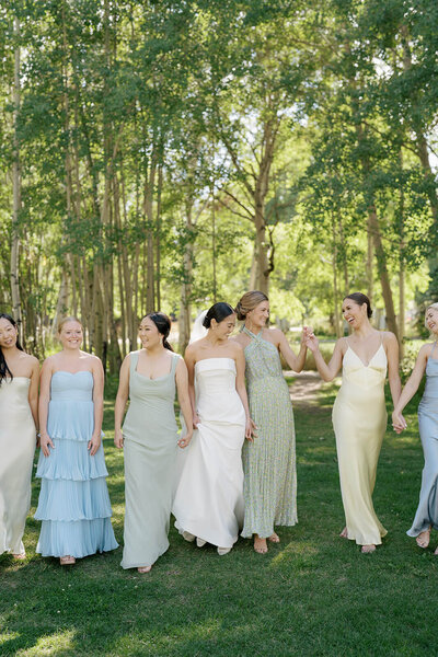 A bride and a her five bridesmaids, who wear dresses in a palette of pale green, blue,  and yellow, walk together through an aspen grove.