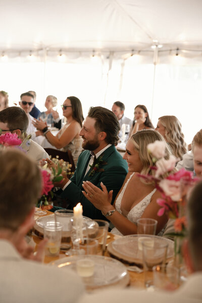 A newlywed couple sits at their wedding reception under a tent, clapping and smiling. The groom wears a teal suit, and the bride is in a lace gown. Guests surround them, enjoying the celebration.