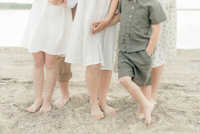 three children stand in front of their parents digging their toes into a Kansas City beach while they hold their siblings' hands. 