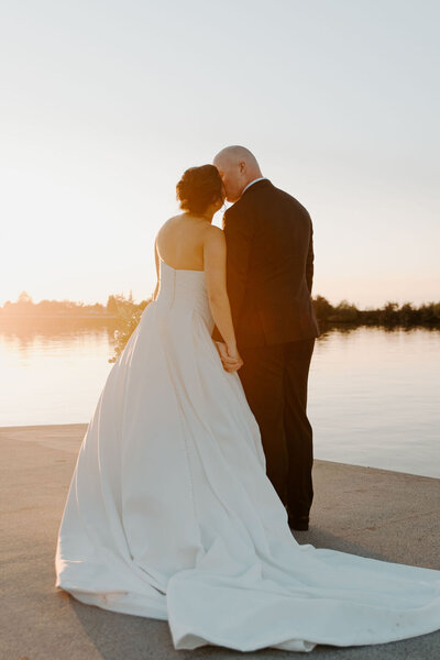 Bride and groom standing by the water at sunset.