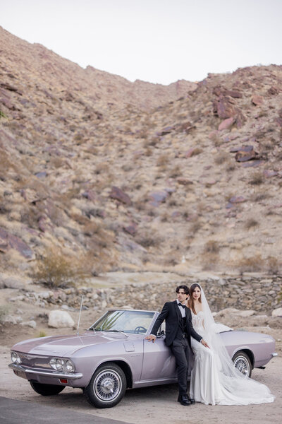 A stylish bride and groom pose beside a vintage convertible in the rugged desert landscape of Palm Springs, California. The couple’s elegant attire contrasts beautifully with the raw, natural surroundings, creating a cinematic and timeless wedding portrait with an old Hollywood feel.