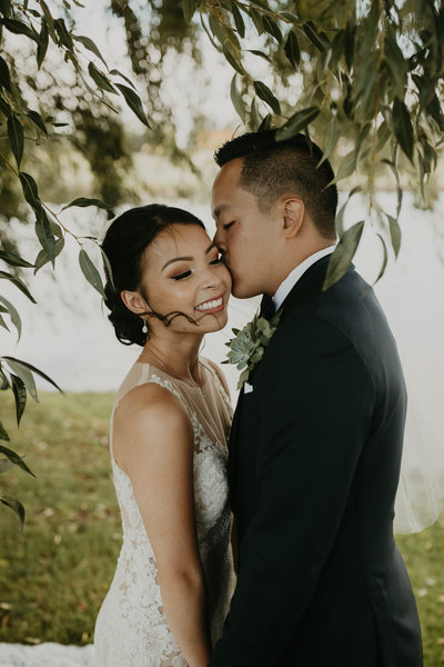 Groom kisses bride's cheek under trees on their wedding day at Langdon Farms in Aurora, Oregon