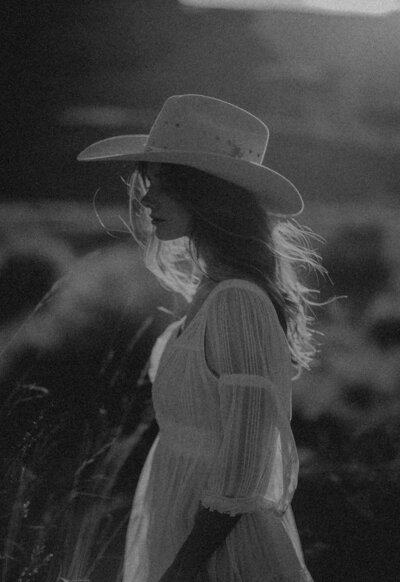 A woman in a white dress and wide-brimmed hat stands outdoors in profile, sunlight highlighting her hair in this black-and-white Sweet Pea Ink Creative photo.