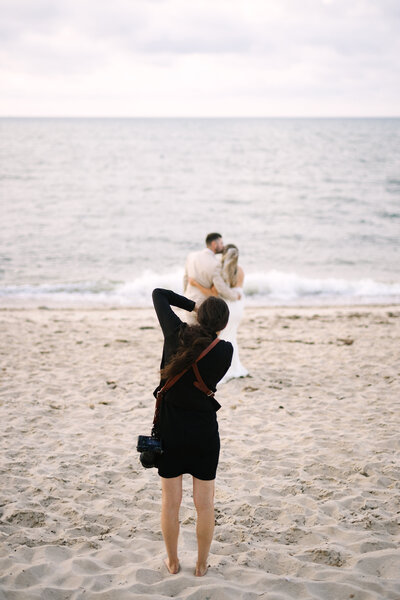image taken from behind cape cod photographer meghan lynch taking a photo of a bride and groom on a beach during their brewster ma wedding