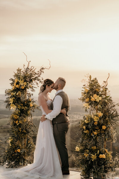 Ein Hochzeitspaar steht eng umschlungen in einen Stirnkuss vor einer malerischen Kulisse in der Toskana umgeben von Blumenranken.