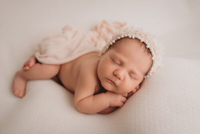 Alpharetta, Georgia newborn photography session with Atlanta's best newborn photographer, Maternity and Newborn Studio of Atlanta by Casey McMinn Photography, capturing a sweet newborn baby girl asleep with her hands under cheek lying on light color fabric and wearing a bonnet.