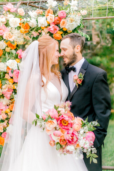 A bride and groom nuzzling in front of a very colorful flower arch