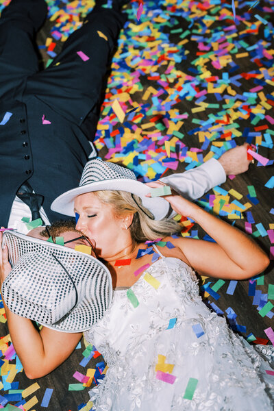 Flash photo of bride and groom in sparkly cowboy hats laying on the ground kissing surrounded by colorful confetti