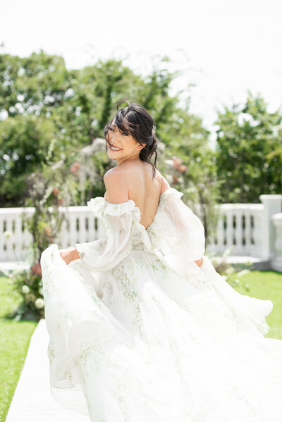 Asian bride in flowing, floral Monique L'Huillier wedding dress runs across terrace and looks back at photographer smiling.