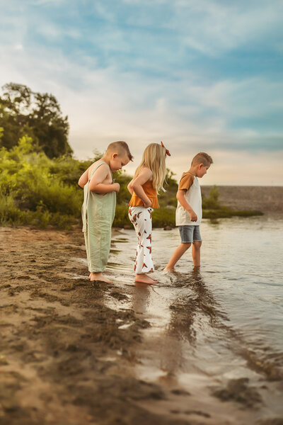 Three barefoot siblings walk into a lake at the beach during sunset summer session