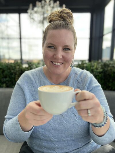 Sarah smiling and holding up a cup of frothy coffee