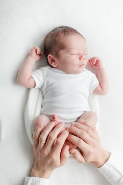 A baby wearing a white onesie and wrapped in a swaddle sleeps while his dad holds his feet.