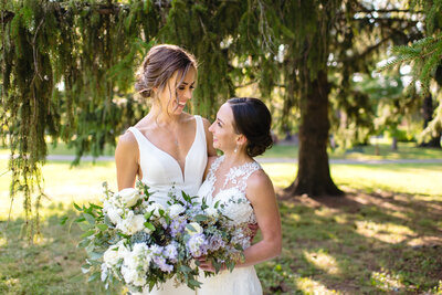 Two brides look into each other's eyes for formal portrait by a willow tree.