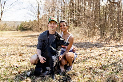 A man and woman kneel on grass in a sunlit clearing with bare trees behind them, joyfully smiling at the camera. They are joined by a black dog with white markings, capturing a moment that any New England family photographer would treasure.