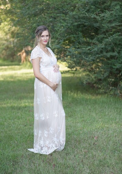 Pregnant mother in a flowing white lace gown stands in a sunlit field, hands gently on her belly. A timeless maternity session by Moments in Light Photography.
