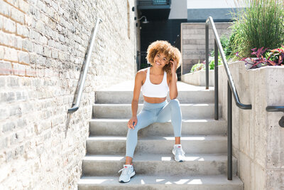 wellness and fitness coach sitting on concrete stairs outdoors, wearing baby blue yoga pants and a white sports crop top