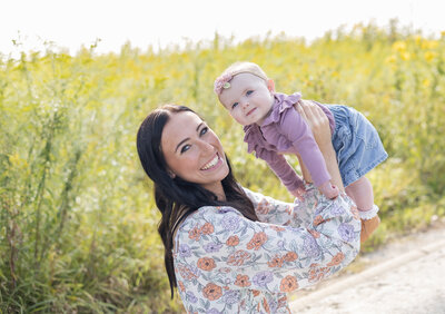 Mom sitting at a wildlife sanctuary with long dark hair, looking at the camera smiling, while holding up her 6 mo old daughter.  Daughter is looking off camera.