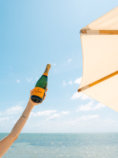 Arm holding champagne bottle in the air next to a beach umbrella