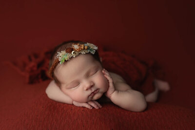 A newborn baby in a floral headband sleeps on a red bed with a hand on her cheek