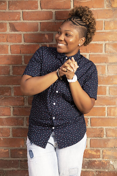 Woman in blue button up top, stonewash blue jeans standing in front of a brick wall