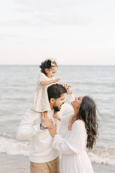 A mother in a white dress sits on a couch tickling and snuggling with her two toddler daughters