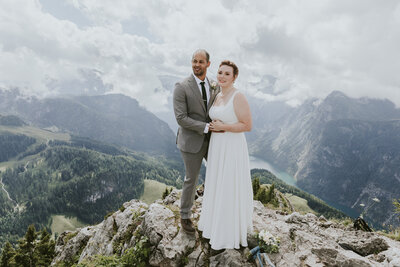 An eloping couple wearing a red dress and grey suit looks out  over Eibsee near Garmisch