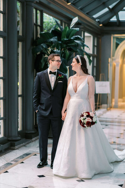 Bride in a white dress carries red and white flowers and walks down the tiled hallway of a DC hotel with her new husband after their wedding in an image by DMV photographer