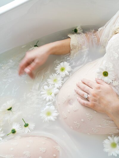 A close-up view of a pregnant belly in a bath surrounded by daisies during a maternity bath photo shoot.
