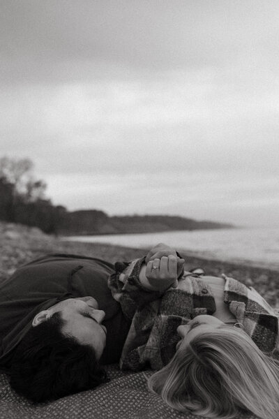 Serene black-and-white image of a couple lying on a quiet beach, holding hands as they gaze at the ocean, reflecting their peaceful, deep bond