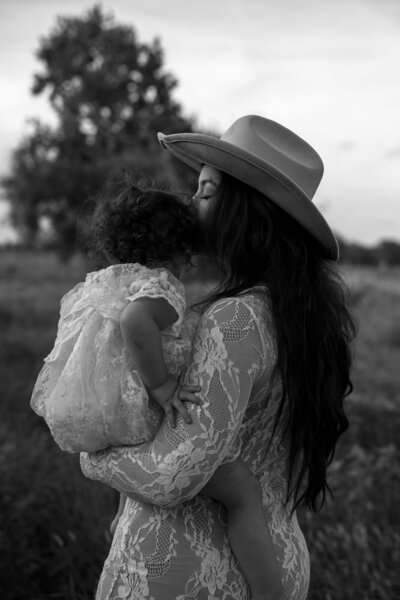A woman in a lace dress and hat tenderly embraces a toddler, sharing a sweet moment in a natural, grassy setting.