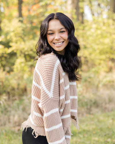 high school senior female with long black hair, turned to the side smiling at the camera, standing in grass with trees behind her, wearing a tan and white stripped shirt.