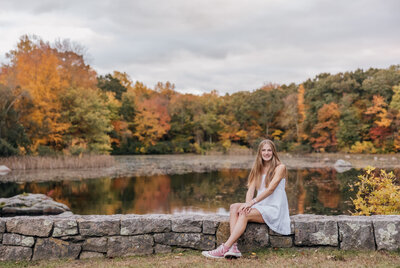high school senior girl sitting on rock wall in front of lake at connecticut college arboretum with fall foliage