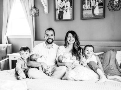 A black and white photo of Reg and Kala holding each holding 1 of their newborn twins. Their other 2 boys are sitting at each of their sides. All of them are smiling and sitting on a bed inside their house.