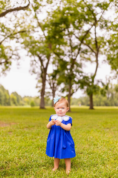 Little girl standing in a field in a blue dress and bow playing with grass
