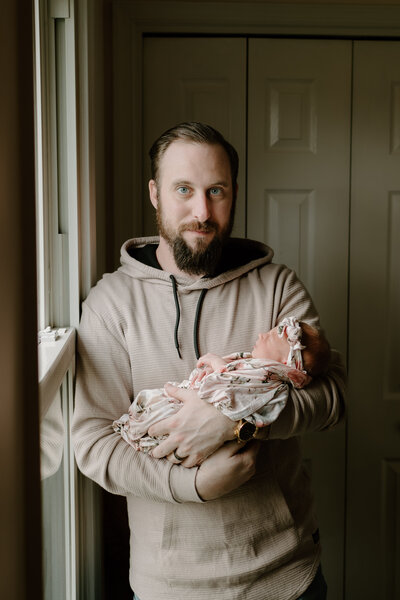 Dad wearing tan standing next to a window in baby girls nursery while holding baby.