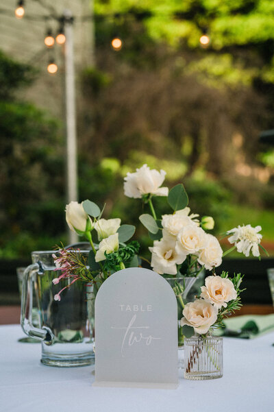 Elegant wedding table centerpiece with white roses, greenery, and a modern table number card