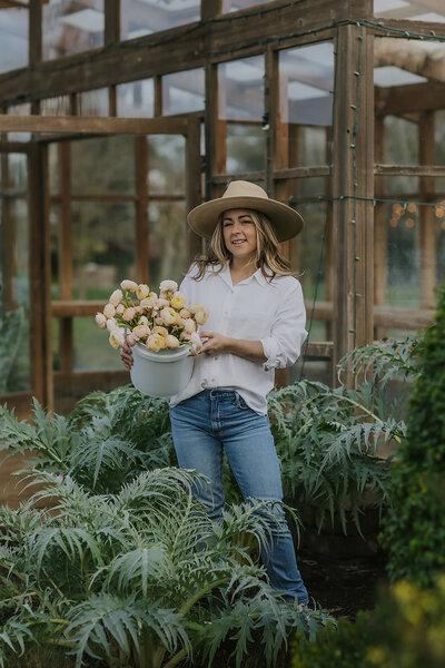 Owner of Peaches and Poppies Floral holding a bucket of flowers and standing in ferns outside of her greenhouse
