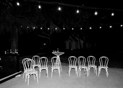 Black and white image of empty row of chairs outside under string lights