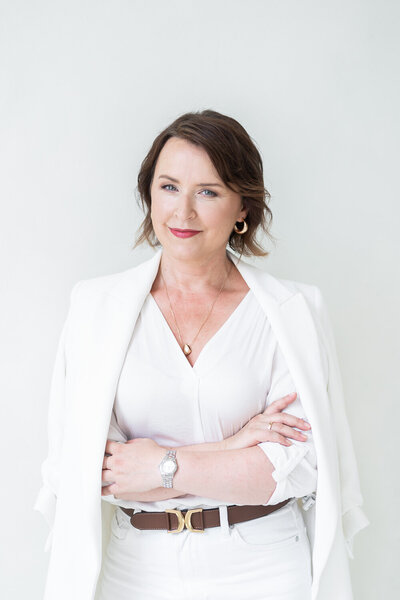 žena s tmavými vlasy v bílém kostýmku se založenýma rukama se dívá s úsměvem do objektivu