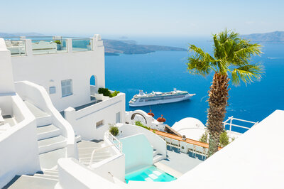 View of white buildings overlooking the sea in Santorini. A cruise ship is sailing on the blue water, with a palm tree in the foreground. Steps and a pool are visible, with a clear, sunny sky above.